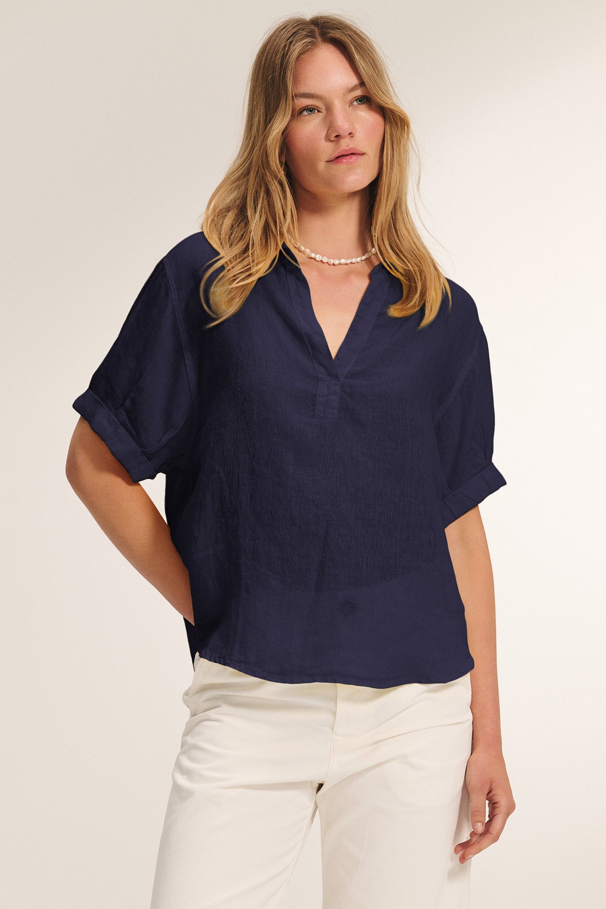 A woman wearing the VIVICA LINEN TOP by Velvet by Graham & Spencer, a navy blue lightweight linen short-sleeve blouse with a V-neckline, paired with white pants, stands against a plain light background.-39292499689665