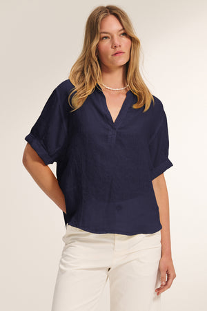 A woman wearing the VIVICA LINEN TOP by Velvet by Graham & Spencer, a navy blue lightweight linen short-sleeve blouse with a V-neckline, paired with white pants, stands against a plain light background.