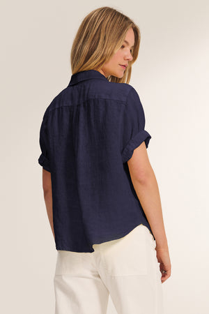 A person with light brown hair stands with their back to the camera against a neutral background, wearing the VIVICA LINEN TOP by Velvet by Graham & Spencer in navy and white pants—a perfect choice for warm-weather style.