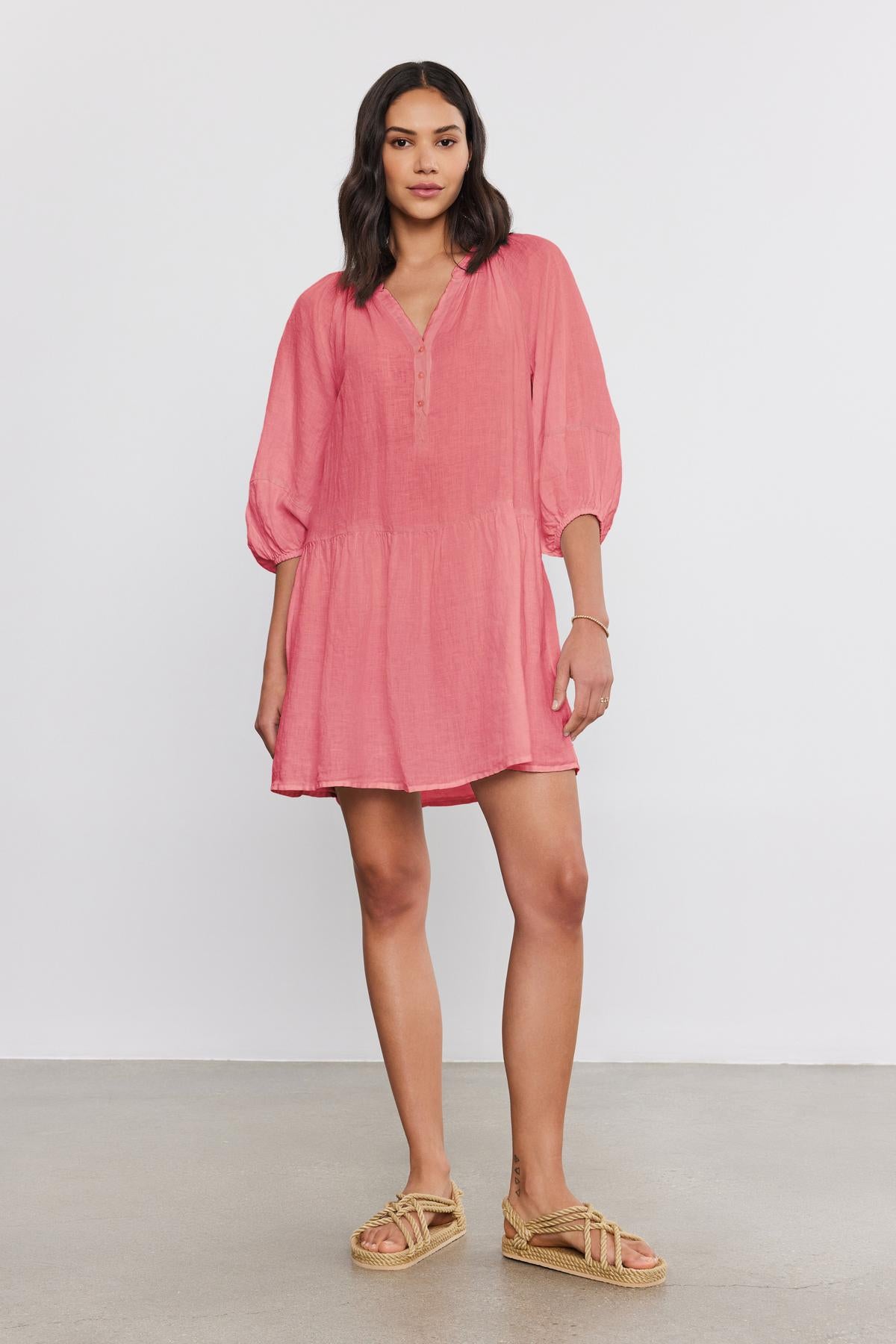   A woman stands against a plain background wearing the VONNIE LINEN DRESS by Velvet by Graham & Spencer, a loose, pink woven linen dress with three-quarter length puff sleeves, paired with tan woven sandals. 