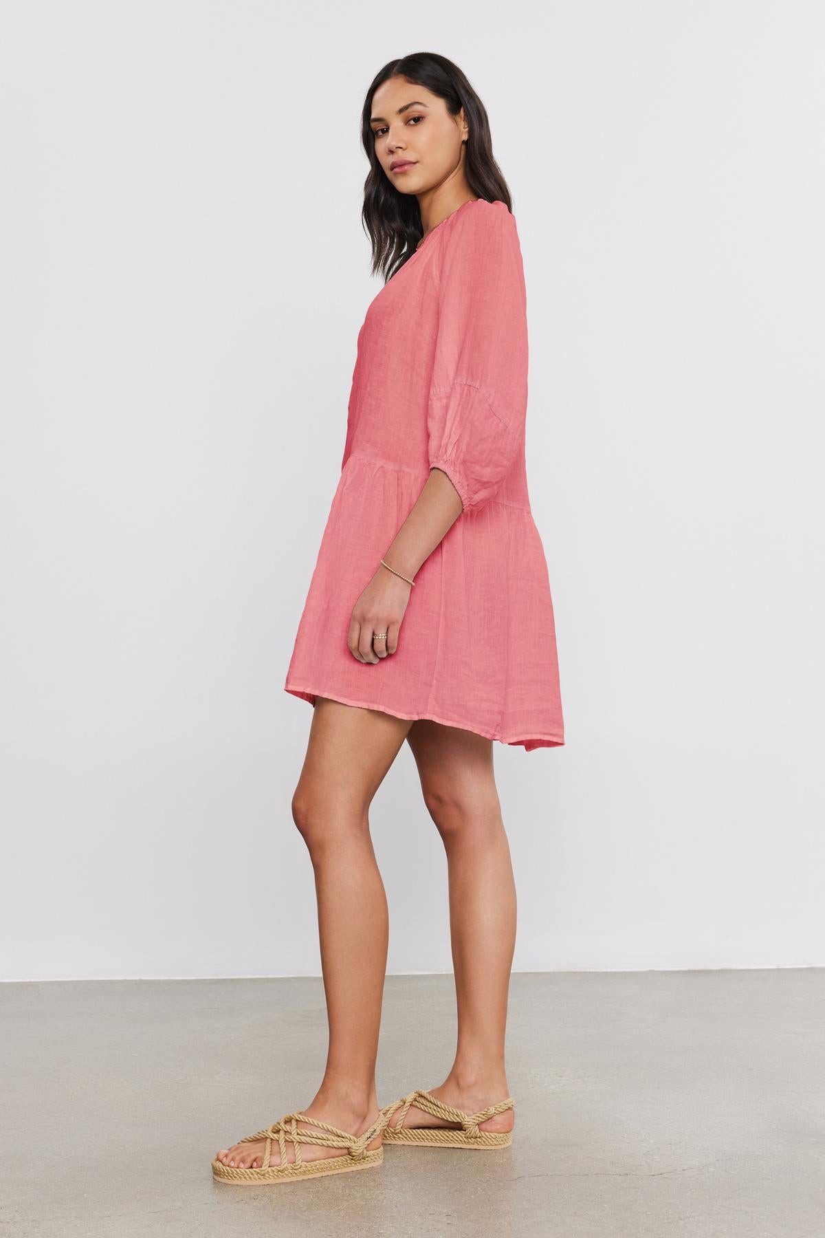   A woman stands sideways against a plain white background, wearing the VONNIE LINEN DRESS by Velvet by Graham & Spencer, paired with beige woven sandals. 