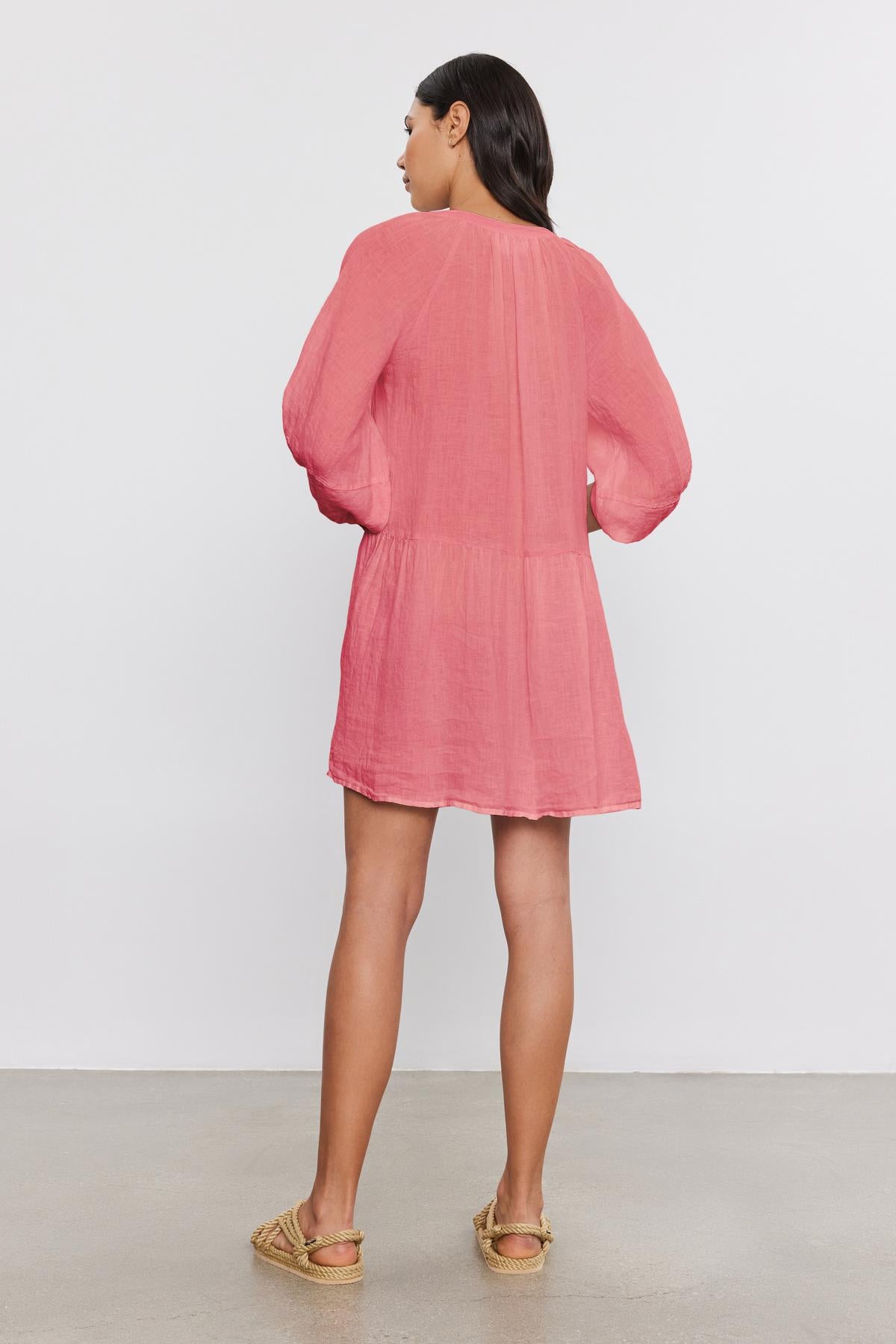   A person stands facing away, wearing the VONNIE LINEN DRESS by Velvet by Graham & Spencer, a pink woven linen dress with puff sleeves, paired with beige slip-on shoes. 