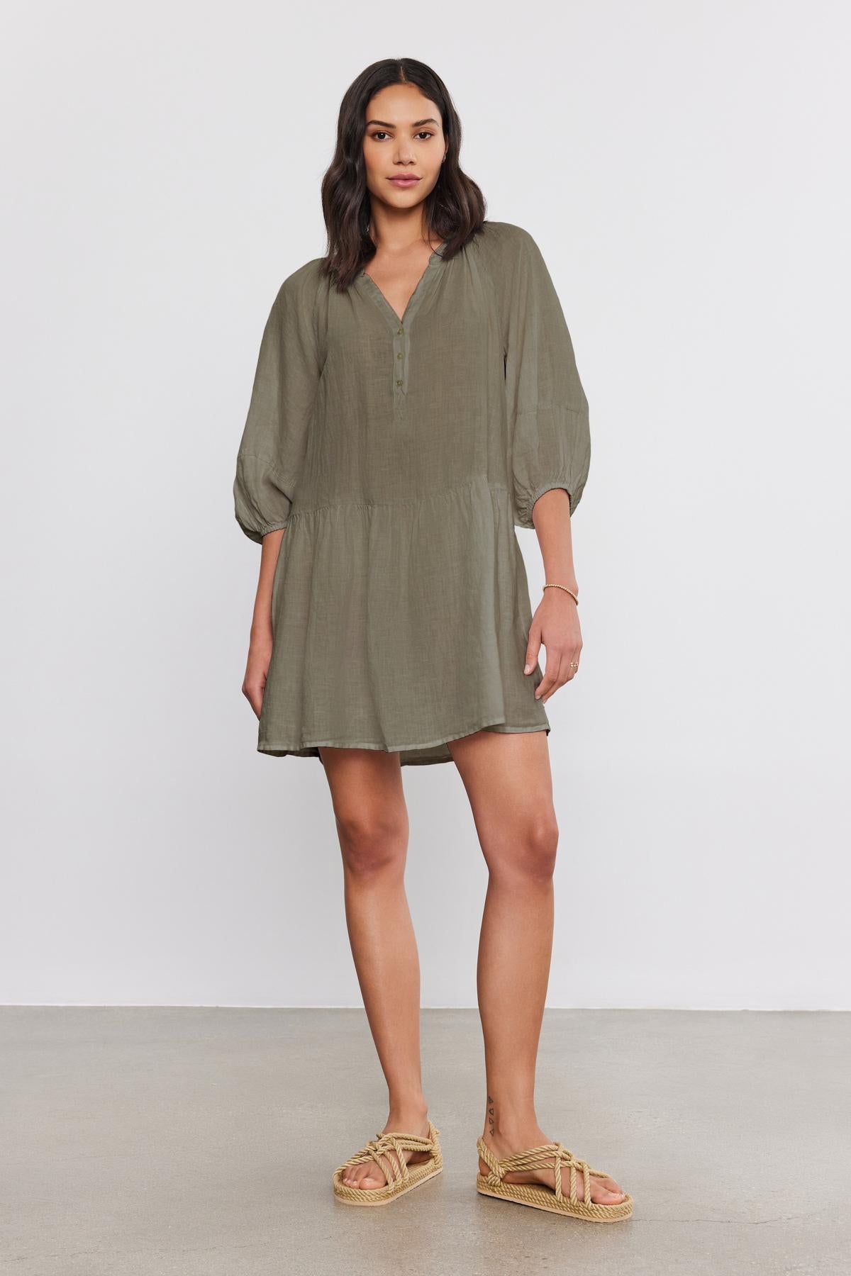   A woman stands against a plain background, wearing the VONNIE LINEN DRESS in light olive green with a split neck and three-quarter puff sleeves by Velvet by Graham & Spencer, paired with beige sandals. 