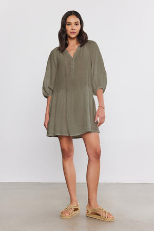 A woman stands against a plain background, wearing the VONNIE LINEN DRESS in light olive green with a split neck and three-quarter puff sleeves by Velvet by Graham & Spencer, paired with beige sandals.