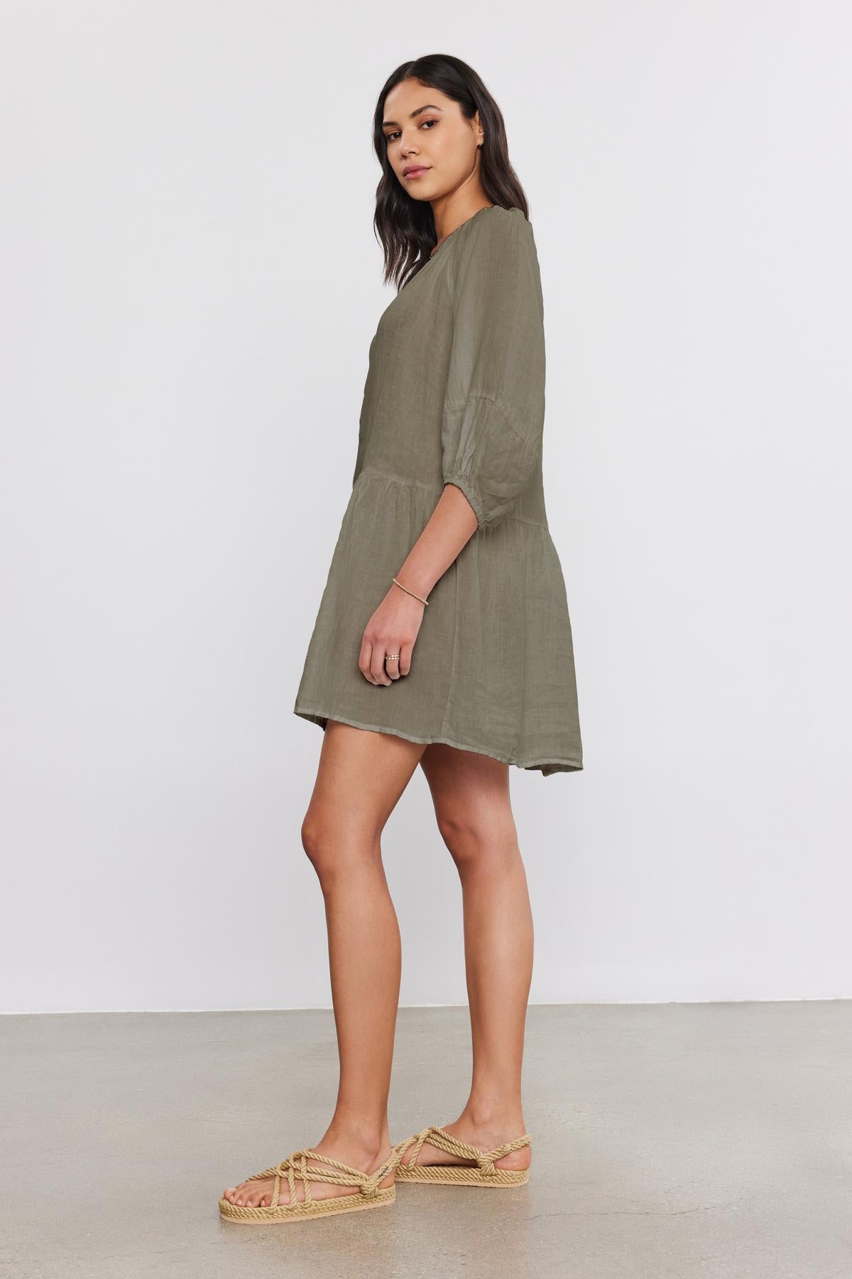   A woman standing and facing slightly to the left, wearing the VONNIE LINEN DRESS by Velvet by Graham & Spencer, an elegant olive green woven linen dress with three-quarter puff sleeves and a split neck. She pairs it with beige woven sandals. She has long dark hair. 