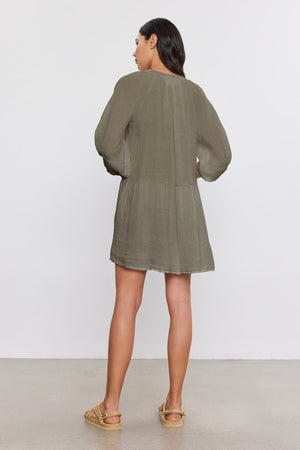 A woman stands facing away from the camera, wearing the VONNIE LINEN DRESS from Velvet by Graham & Spencer. The dress features a loose-fitting design with a split neck and puff sleeves. Her tan espadrille shoes complement the outfit as she stands against a plain white background.