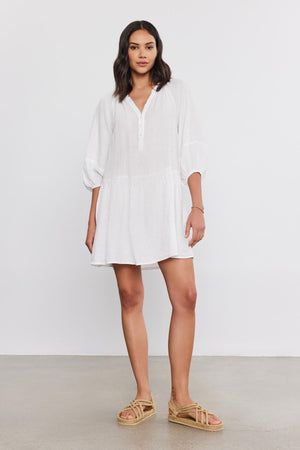 A woman stands in a studio, wearing a white VONNIE LINEN DRESS from Velvet by Graham & Spencer with a split neck and straw sandals, looking at the camera with a neutral expression.