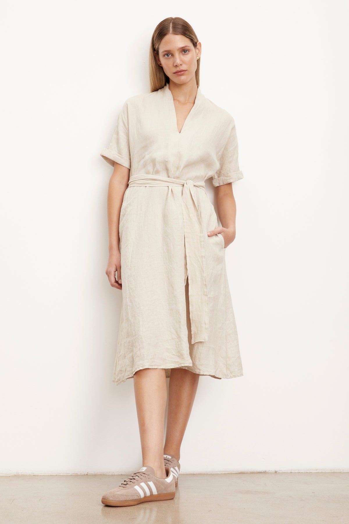 Woman leaning against wall wearing Winley Dress in sand colored linen belted with hand in pocket full length front-26426151993537
