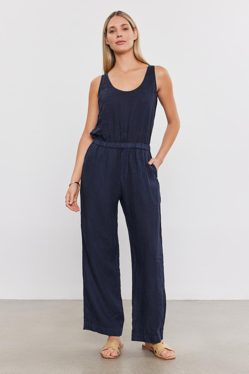 A woman in a dark blue Velvet by Graham & Spencer Winnie Linen Jumpsuit stands against a plain background, looking directly at the camera with her hands at her sides.