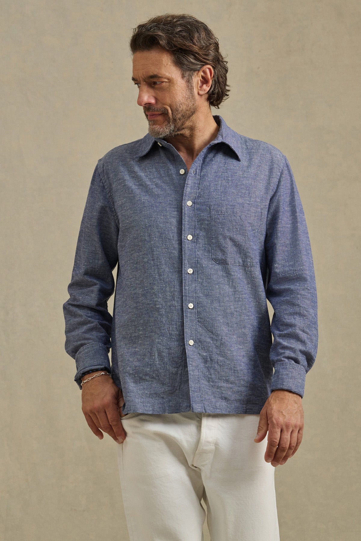 Man wearing the ANTOINE SHIRT by Velvet by Graham & Spencer—a blue cotton chambray button-up with a classic collar—paired with white pants stands against a plain beige background, looking to his left.