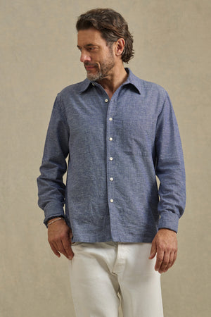 Man wearing the ANTOINE SHIRT by Velvet by Graham & Spencer—a blue cotton chambray button-up with a classic collar—paired with white pants stands against a plain beige background, looking to his left.