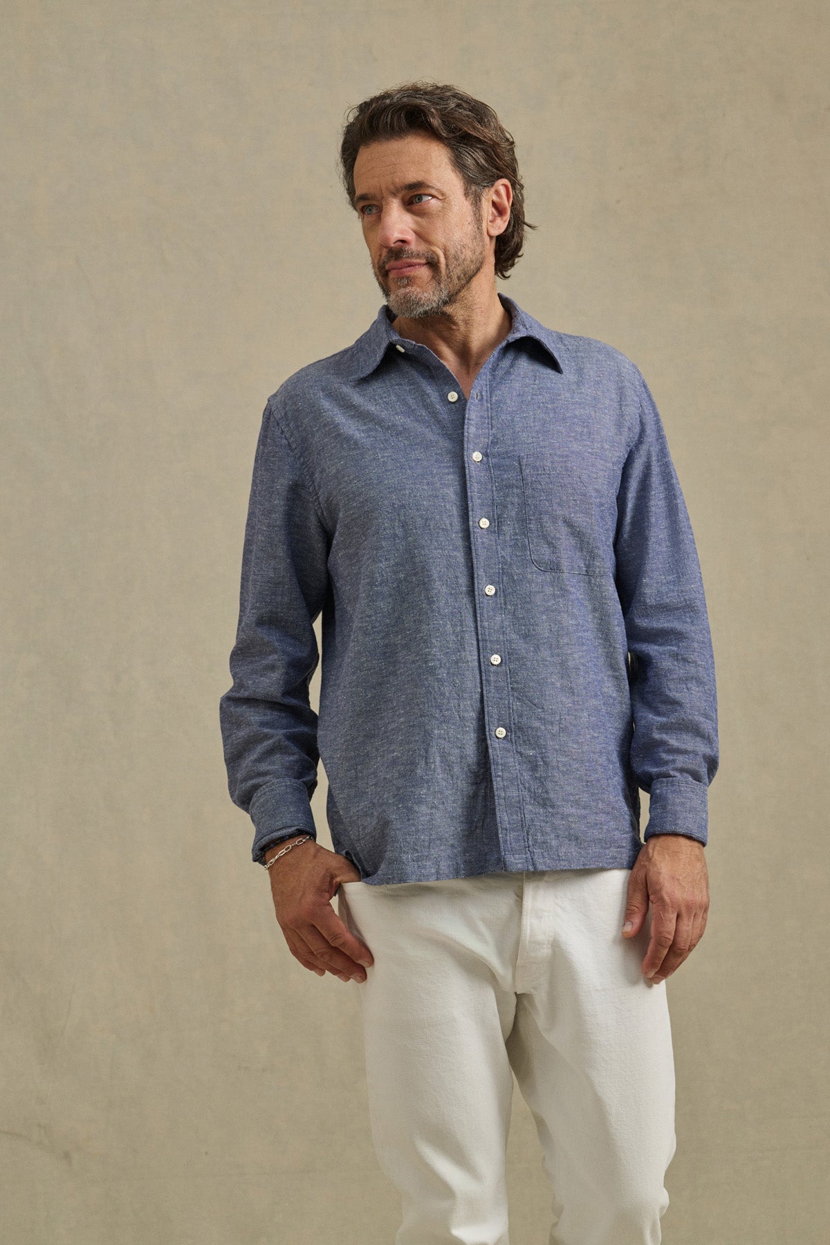 A man stands against a plain background wearing the ANTOINE SHIRT by Velvet by Graham & Spencer—a blue chambray button-up with a classic collar—and white pants, looking slightly to his left.