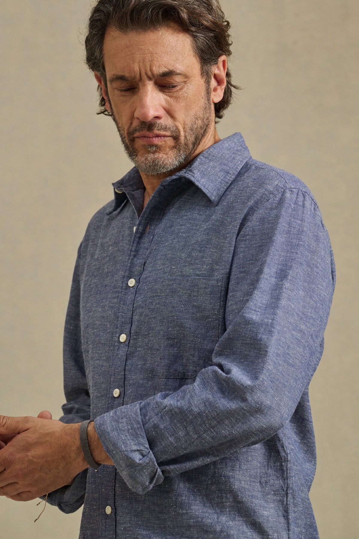 A man with dark hair and a beard wears the ANTOINE SHIRT by Velvet by Graham & Spencer, a blue cotton chambray button-up with rolled sleeves, looking down as he adjusts his cuff.