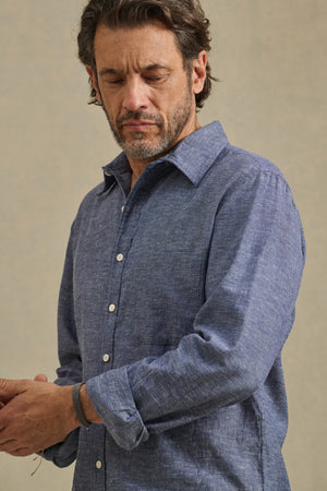 A man with dark hair and a beard wears the ANTOINE SHIRT by Velvet by Graham & Spencer, a blue cotton chambray button-up with rolled sleeves, looking down as he adjusts his cuff.