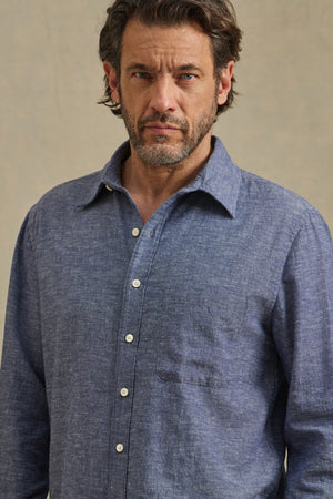 A man with short brown hair and a beard wears the ANTOINE SHIRT by Velvet by Graham & Spencer, a blue chambray button-up with a classic collar, looking directly at the camera against a plain background.