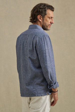 A man with medium-length brown hair, wearing the ANTOINE SHIRT by Velvet by Graham & Spencer in blue and light pants, stands sideways against a neutral background.