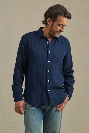 A man wears the APOLLO SHIRT by Velvet by Graham & Spencer, a relaxed navy cotton gauze button-up with a classic collar, paired with light blue jeans. He stands against a plain beige background, looking down to his left.