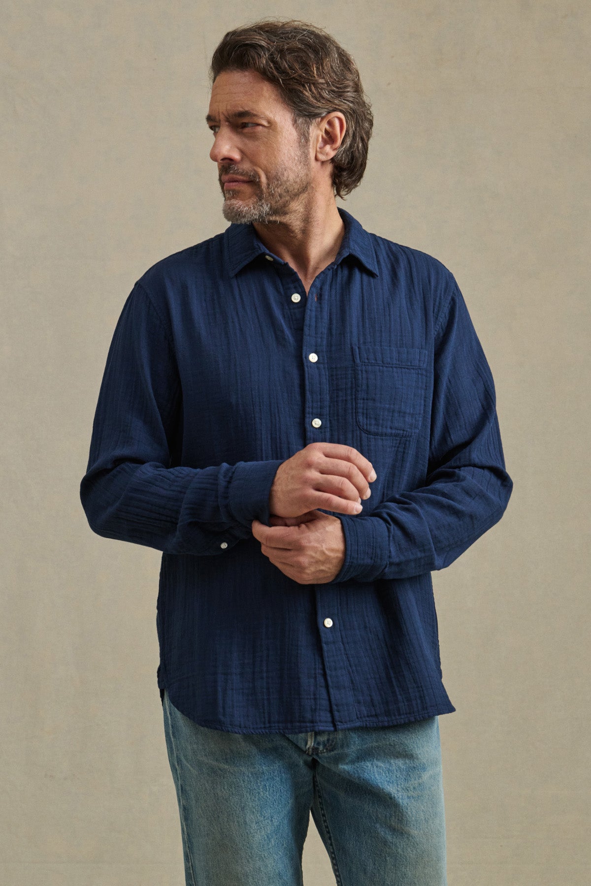 A man with wavy brown hair and a beard wears the APOLLO SHIRT by Velvet by Graham & Spencer, featuring a classic collar, styled with light blue jeans. He stands against a plain beige background, looking to his left.