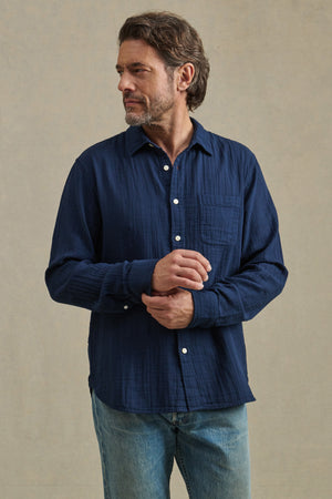 A man with wavy brown hair and a beard wears the APOLLO SHIRT by Velvet by Graham & Spencer, featuring a classic collar, styled with light blue jeans. He stands against a plain beige background, looking to his left.