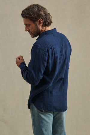 A man with brown hair, seen from behind, wears the APOLLO SHIRT by Velvet by Graham & Spencer and light blue jeans, standing against a plain beige background.