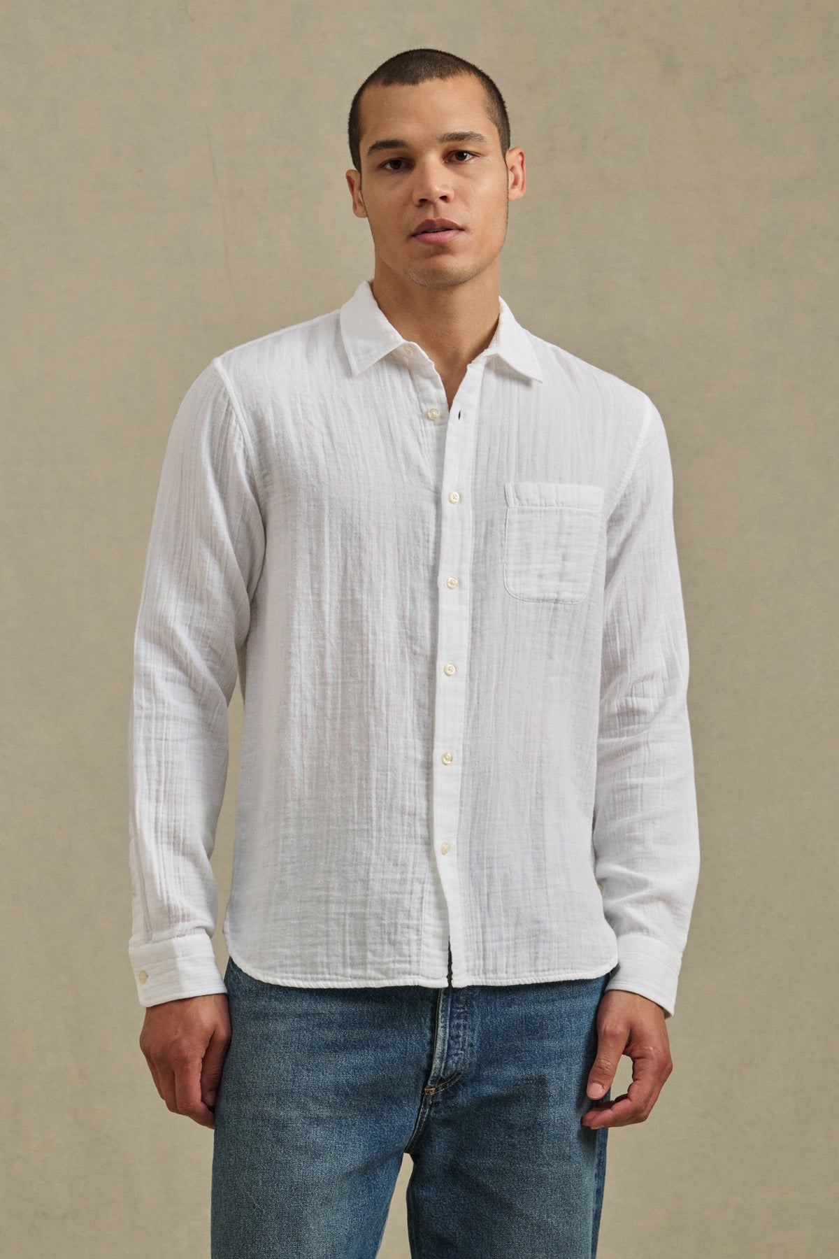 A man stands against a plain beige background wearing Velvet by Graham & Spencer's APOLLO SHIRT, a relaxed cotton gauze style with a classic collar, paired with blue jeans.-41297073995969