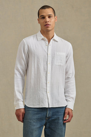 A man stands against a plain beige background wearing Velvet by Graham & Spencer's APOLLO SHIRT, a relaxed cotton gauze style with a classic collar, paired with blue jeans.