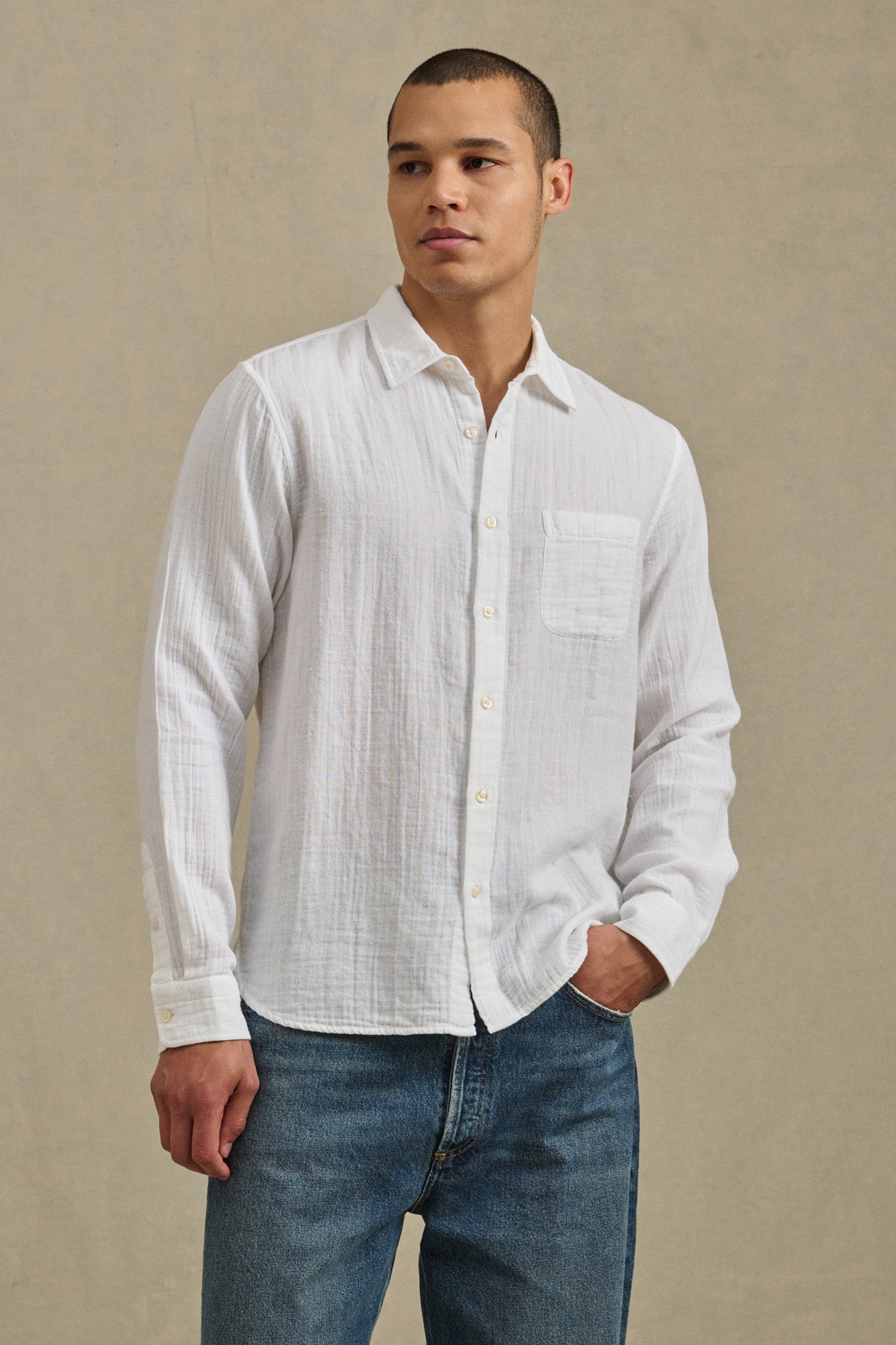 A person wears the APOLLO SHIRT by Velvet by Graham & Spencer—a white relaxed button-up with a front pocket—paired with blue jeans, standing against a plain beige background.-41297073963201