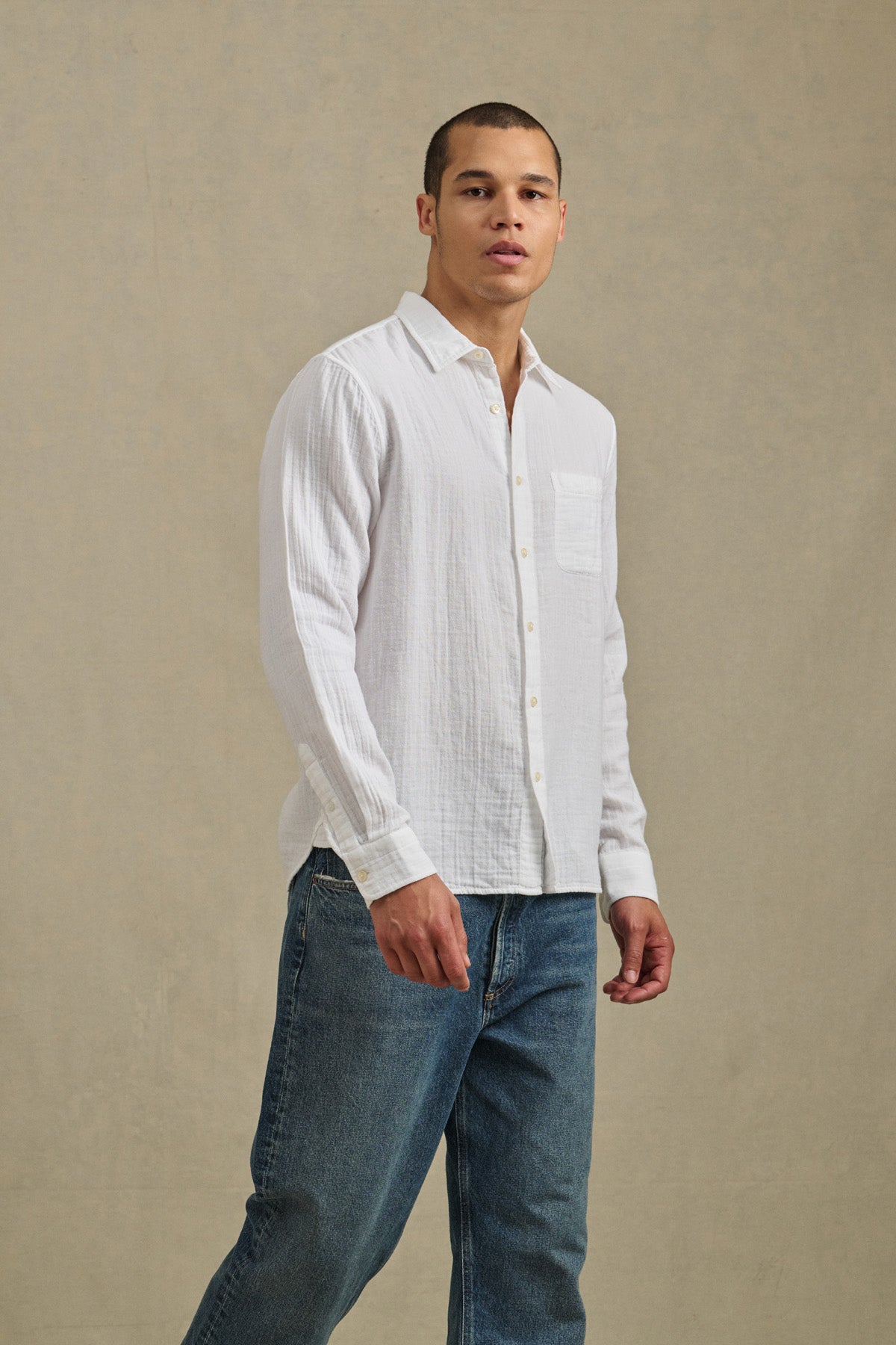 A person stands against a plain beige background, facing the camera in a neutral expression, wearing the APOLLO SHIRT by Velvet by Graham & Spencer—a relaxed button-up in lightweight cotton gauze—paired with blue jeans.