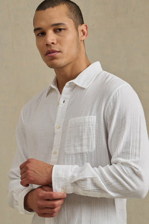 A man wearing the APOLLO SHIRT by Velvet by Graham & Spencer, featuring a relaxed fit, classic collar, and chest pocket, stands against a plain beige background and looks at the camera.