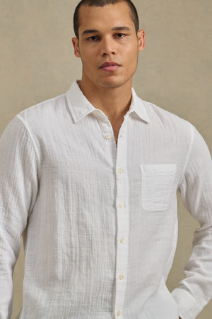 A man wearing the APOLLO SHIRT by Velvet by Graham & Spencer, a white classic-collar button-up crafted from lightweight cotton gauze, stands against a neutral background.