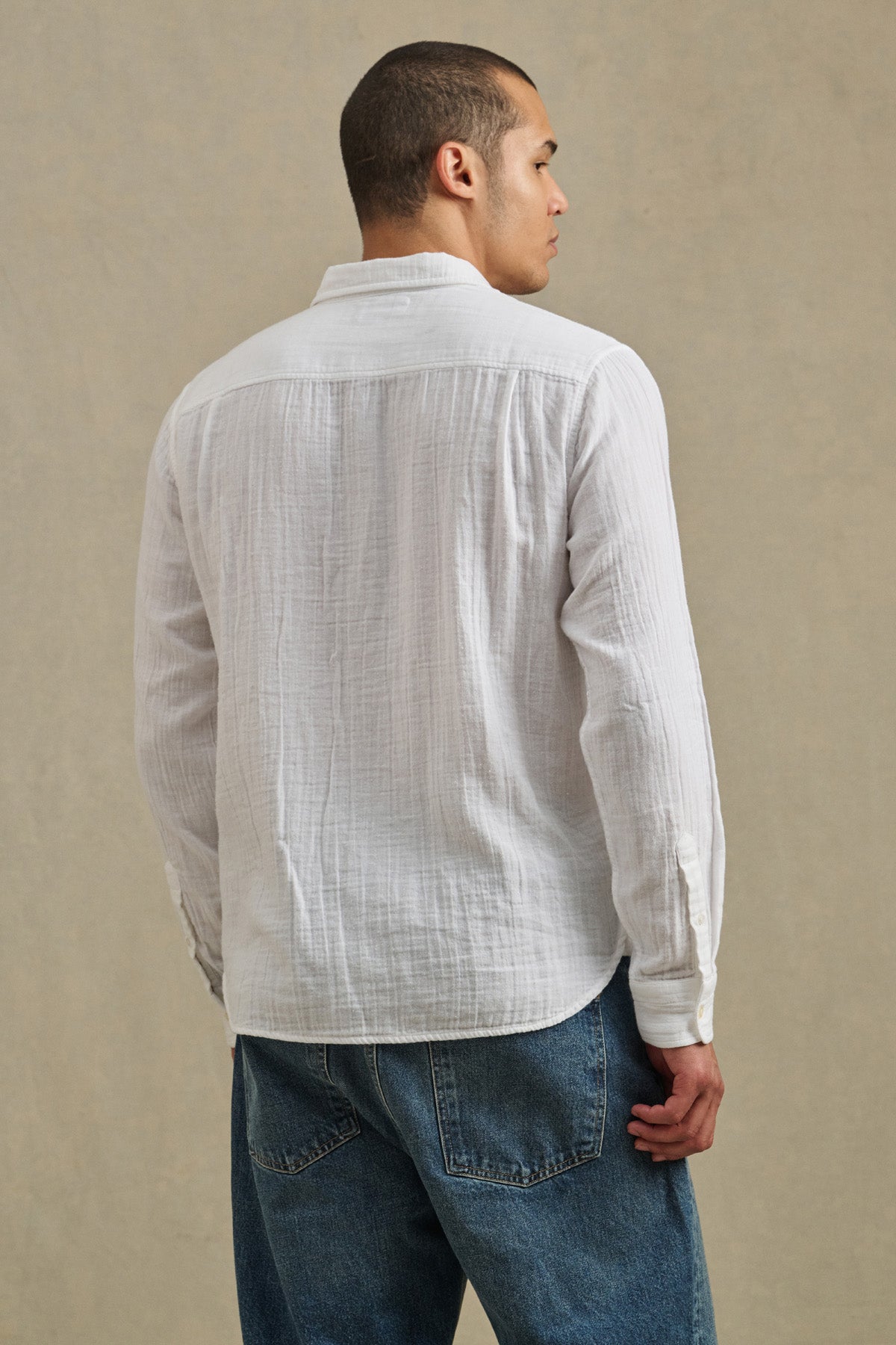 A man wearing the APOLLO SHIRT by Velvet by Graham & Spencer, a relaxed lightweight cotton gauze button-up, and blue jeans stands facing away from the camera against a plain background.