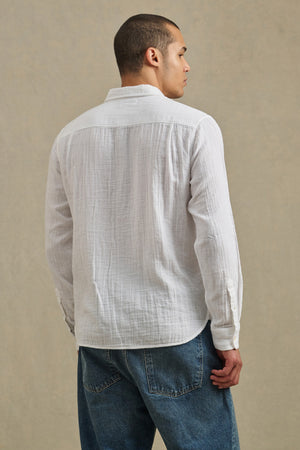 A man wearing the APOLLO SHIRT by Velvet by Graham & Spencer, a relaxed lightweight cotton gauze button-up, and blue jeans stands facing away from the camera against a plain background.