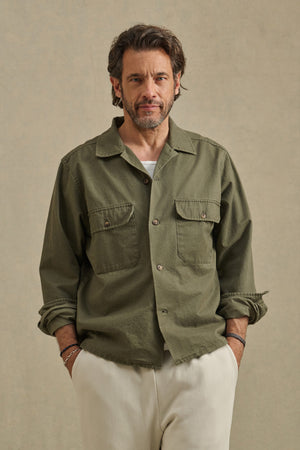 A man with brown hair and a beard is wearing the ARCHER SHIRT by Velvet by Graham & Spencer in olive green over a white T-shirt and cream-colored pants, standing against a neutral background.