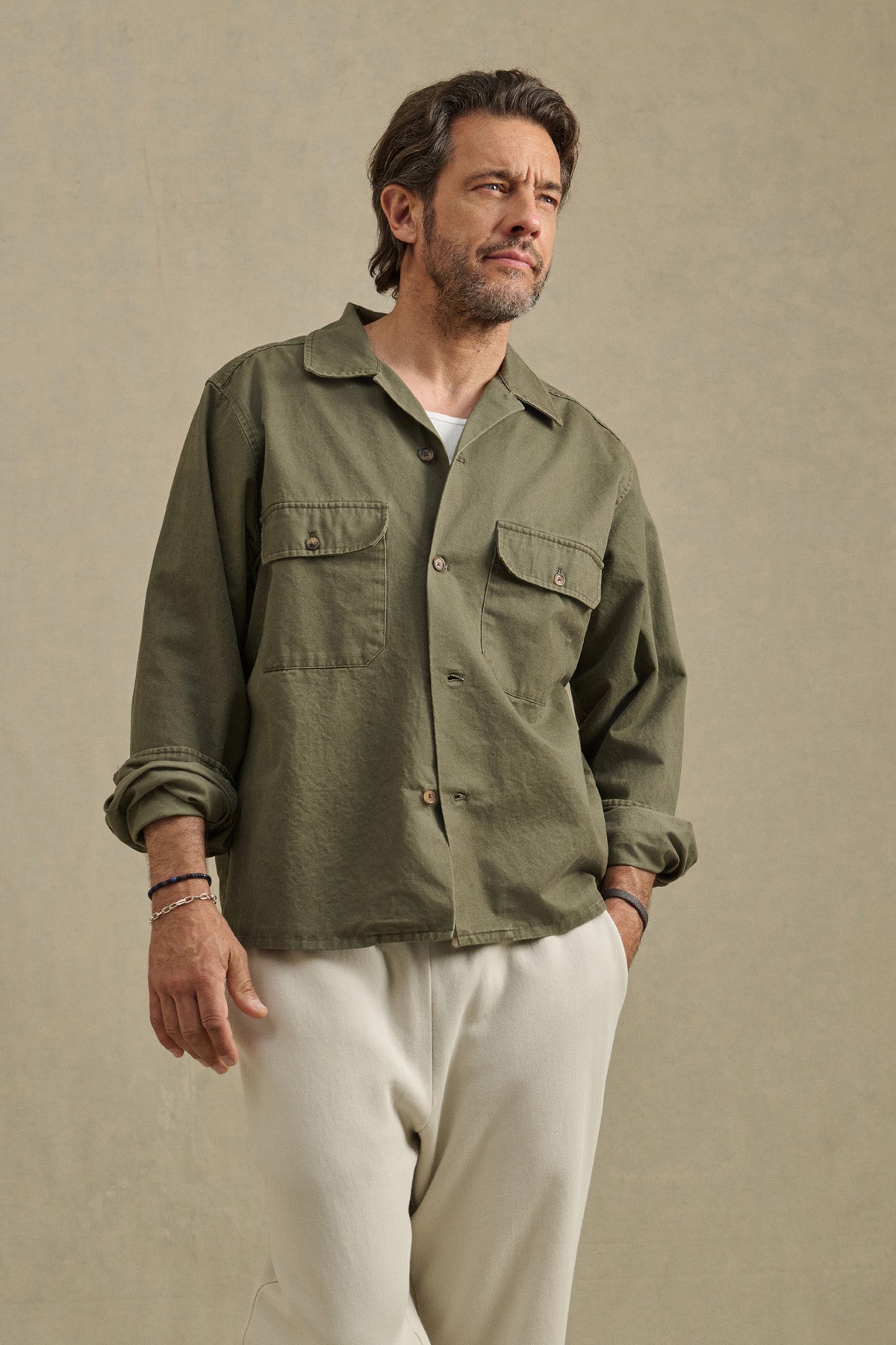 A man stands against a neutral background wearing the ARCHER SHIRT by Velvet by Graham & Spencer in olive green cotton twill with front pockets, paired with off-white pants.-40850642567361