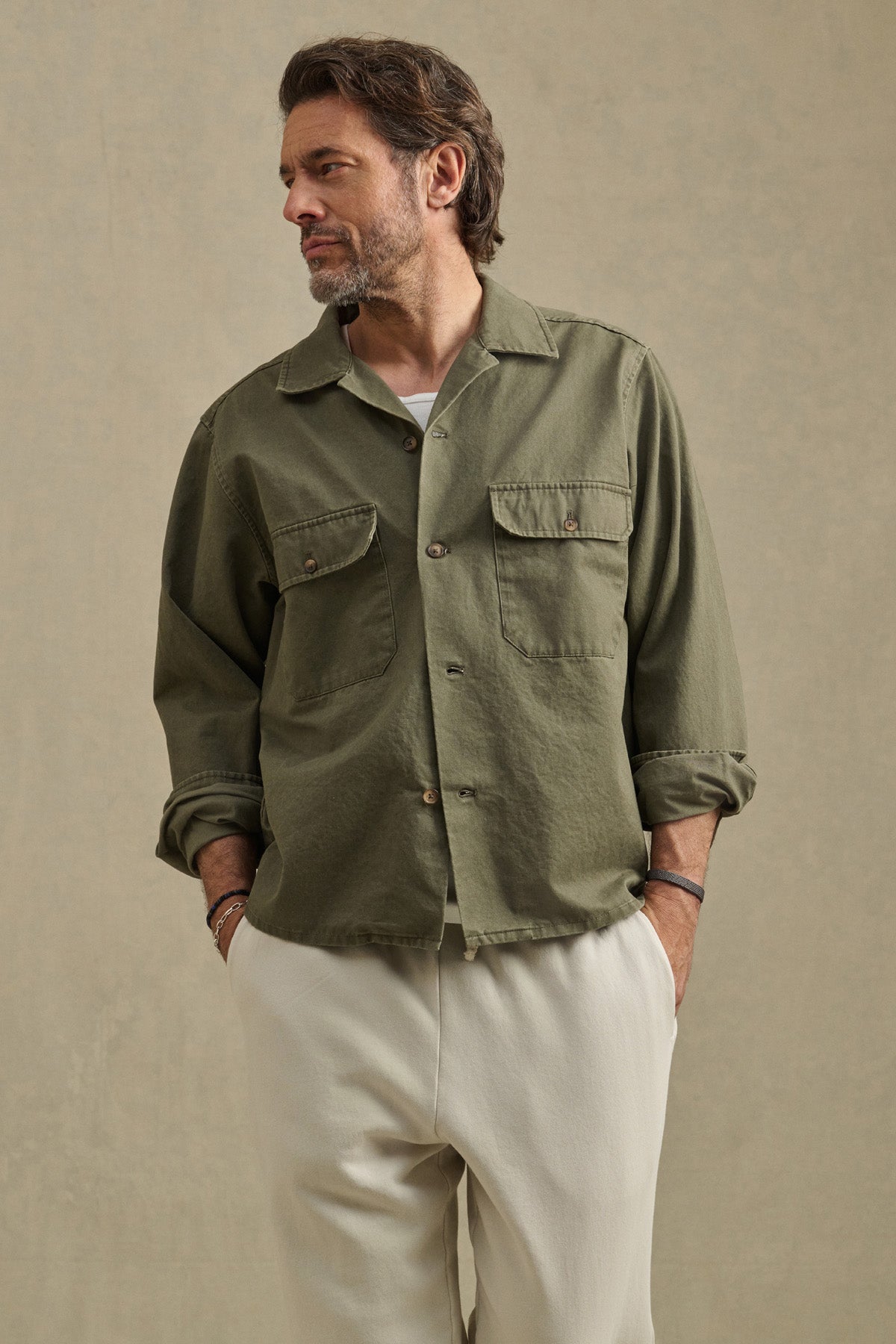 A man stands with hands in pockets, wearing the ARCHER SHIRT by Velvet by Graham & Spencer in green over a white t-shirt and beige pants, looking to his left against a neutral background.