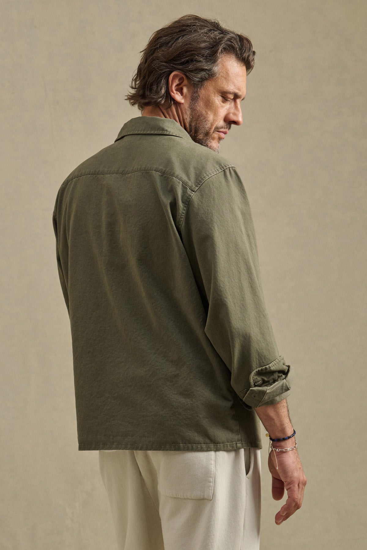 A man stands with his back turned, wearing the ARCHER SHIRT in olive green cotton twill by Velvet by Graham & Spencer, sleeves rolled, paired with off-white pants against a plain beige background.-40850642469057