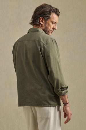 A man stands with his back turned, wearing the ARCHER SHIRT in olive green cotton twill by Velvet by Graham & Spencer, sleeves rolled, paired with off-white pants against a plain beige background.