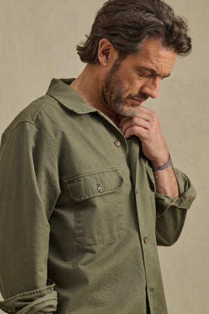 A man with medium-length brown hair and a beard wears the ARCHER SHIRT in olive green cotton twill by Velvet by Graham & Spencer, looking downward and touching his chin against a neutral background.
