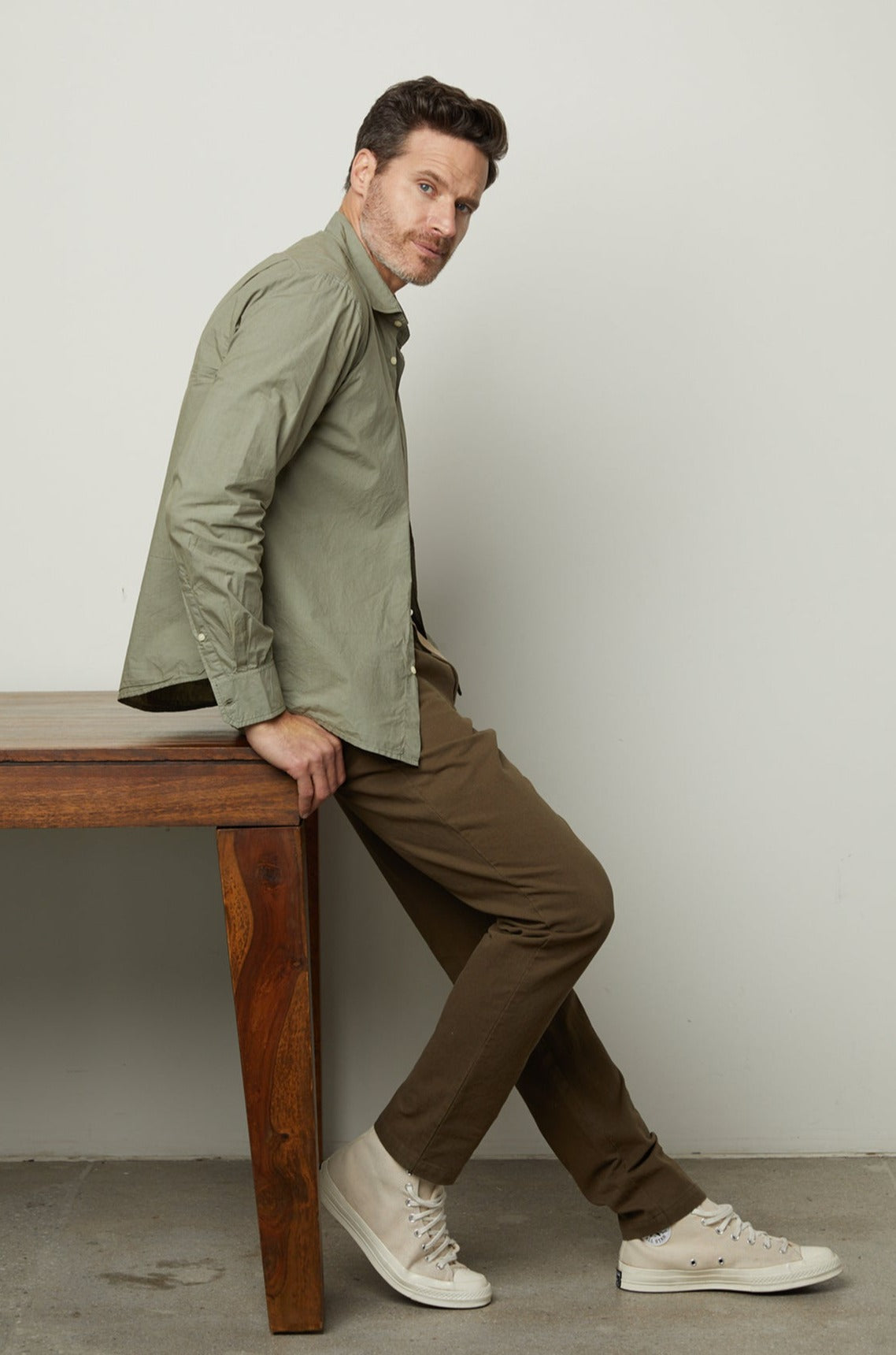   A man leaning against a wooden table in a green shirt wearing the Moran Cotton Twill Pant by Velvet by Graham & Spencer. 