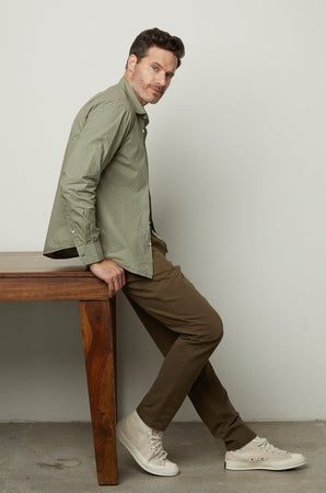 A person leans against a wooden table while wearing a light green shirt, MORAN PANT by Velvet by Graham & Spencer, and white sneakers. The background is plain white.