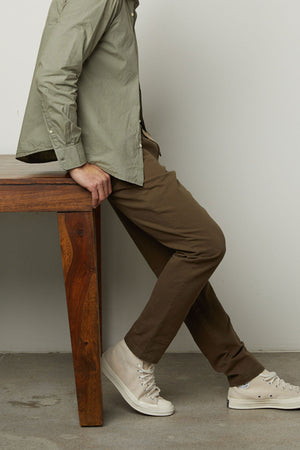 A person wearing a light green shirt, the Velvet by Graham & Spencer MORAN PANT in brown cotton twill, and white sneakers sits on the edge of a wooden table with legs extended and arms resting beside them.