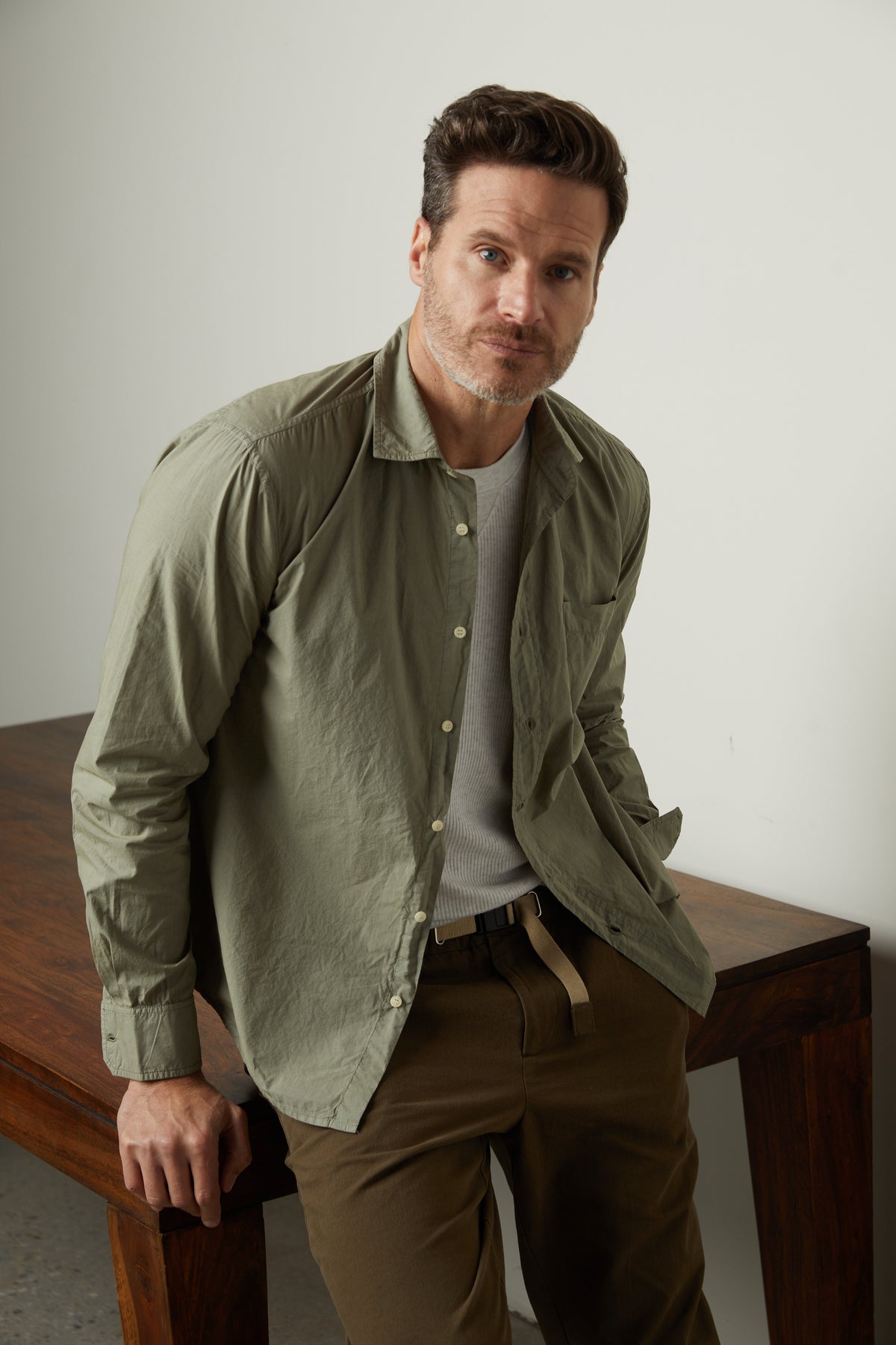   A man in a BROOKS BUTTON-UP SHIRT by Velvet by Graham & Spencer and khaki pants is leaning against a wooden table. 