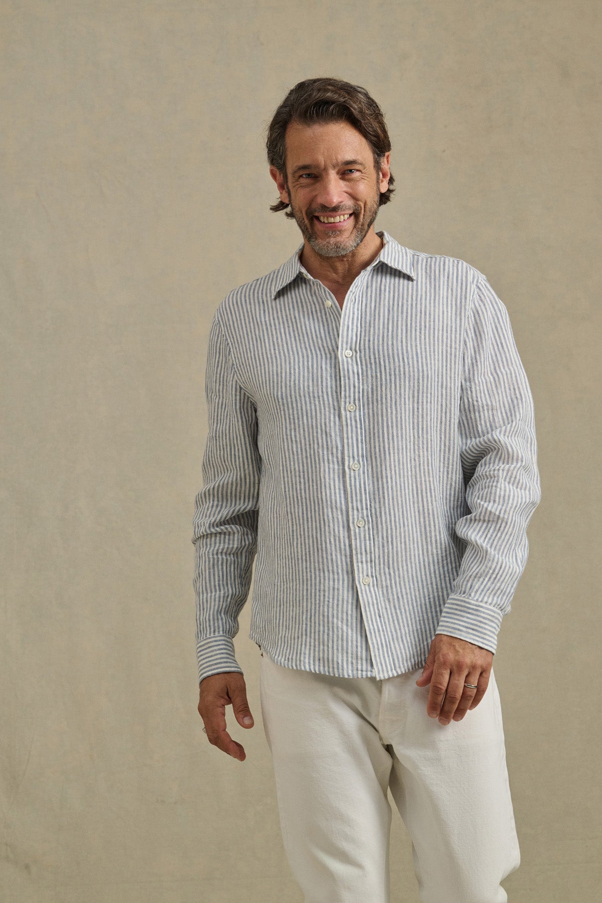 A man smiles as he wears the CARUSO LINEN SHIRT by Velvet by Graham & Spencer—a light blue and white striped button-up—paired with white pants, set against a plain beige background.