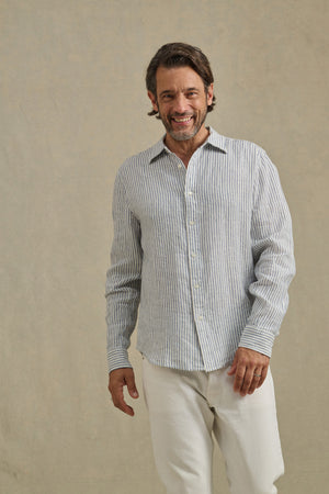 A man smiles as he wears the CARUSO LINEN SHIRT by Velvet by Graham & Spencer—a light blue and white striped button-up—paired with white pants, set against a plain beige background.
