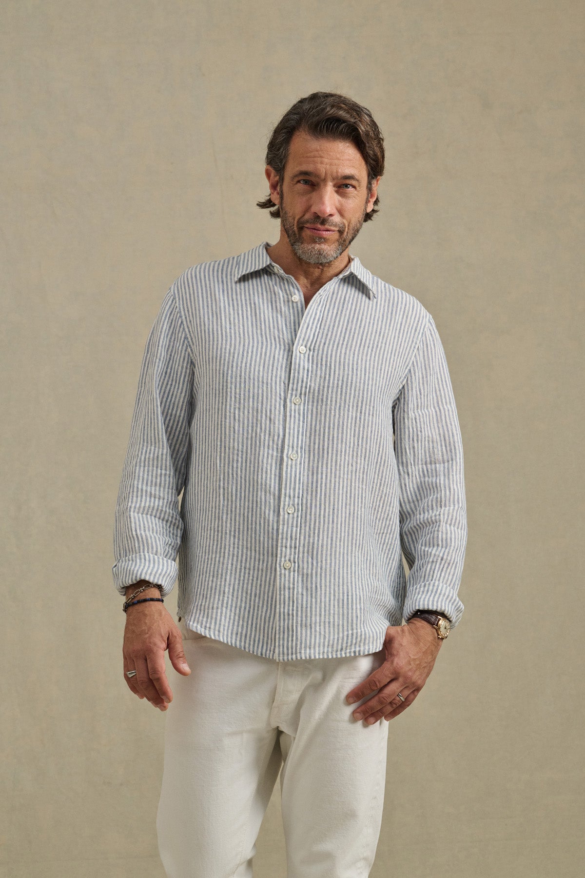 A man wears the CARUSO LINEN SHIRT by Velvet by Graham & Spencer—light blue and white striped—paired with white pants, standing against a plain beige background.