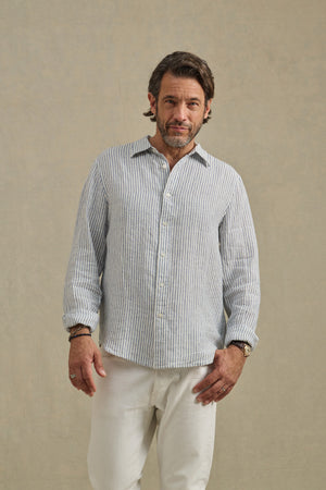 A man wears the CARUSO LINEN SHIRT by Velvet by Graham & Spencer—light blue and white striped—paired with white pants, standing against a plain beige background.