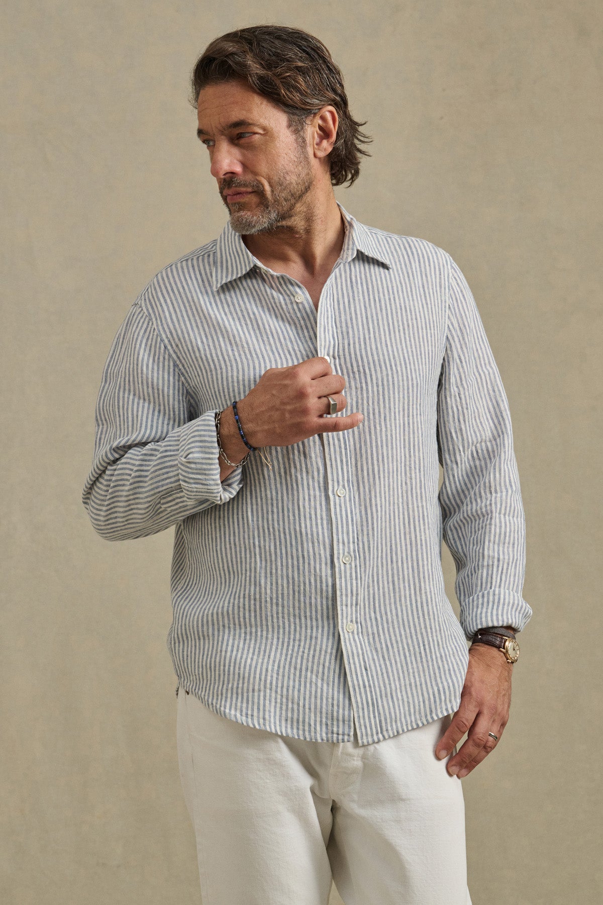 A man wearing the Velvet by Graham & Spencer CARUSO LINEN SHIRT in light blue and white stripes with white pants stands against a neutral background, looking to his left.