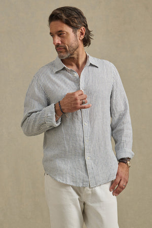 A man wearing the Velvet by Graham & Spencer CARUSO LINEN SHIRT in light blue and white stripes with white pants stands against a neutral background, looking to his left.
