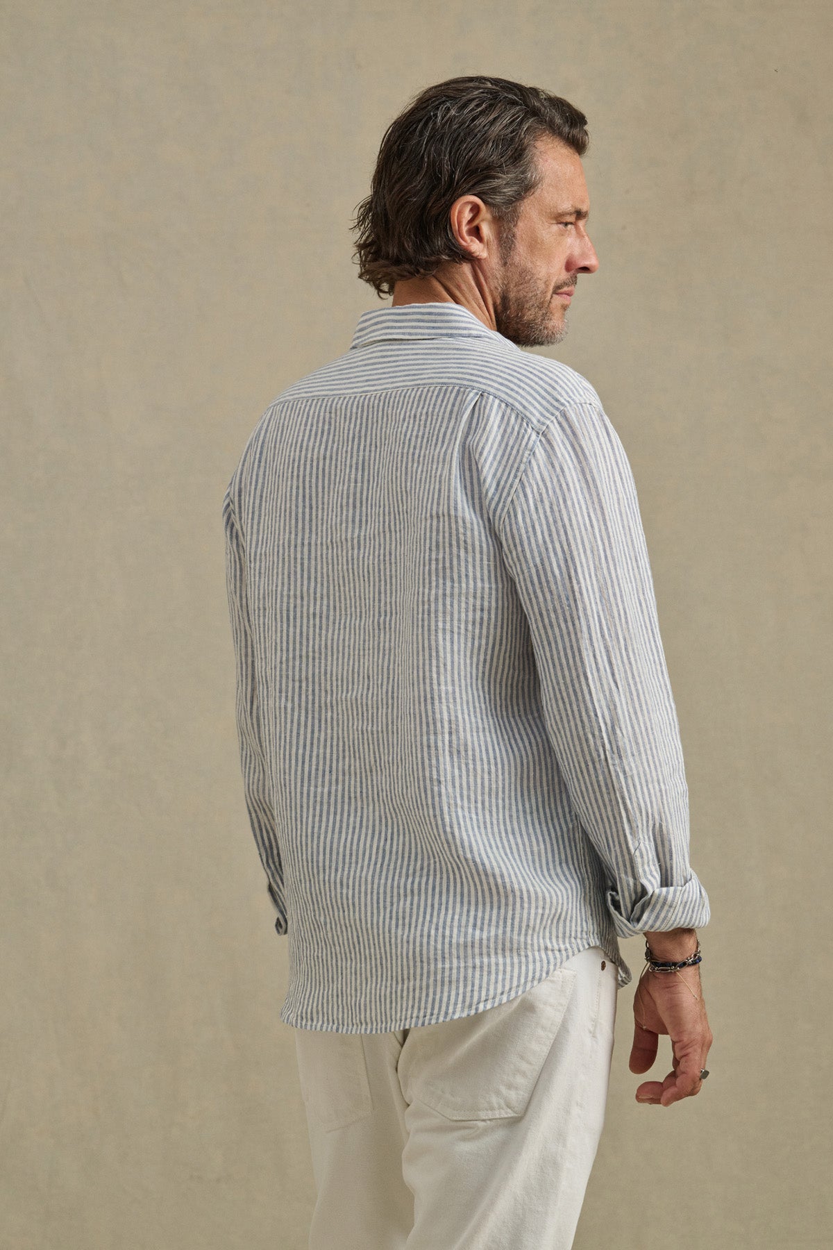 A man with graying hair wears the Velvet by Graham & Spencer CARUSO LINEN SHIRT, a light blue and white striped long-sleeve, and white pants, standing with his back to the camera against a neutral background.-41145256542401