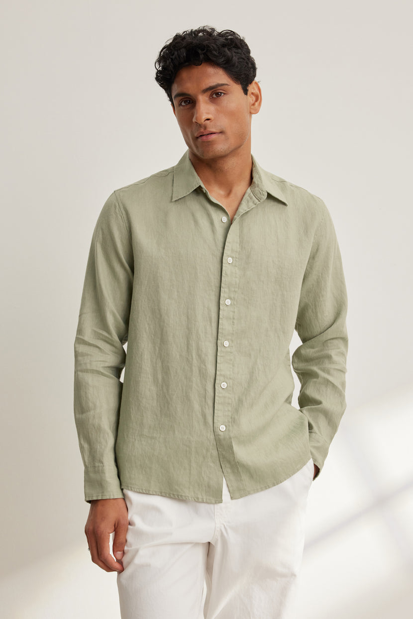 A man wears the CARUSO LINEN BUTTON-UP SHIRT by Velvet by Graham & Spencer in light green with white pants, standing against a plain, light-colored background.